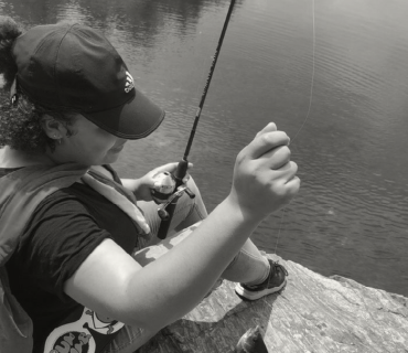 A girl sitting on the edge of a rock with a fishing pole, overlooking a lake, considering computer science outdoors