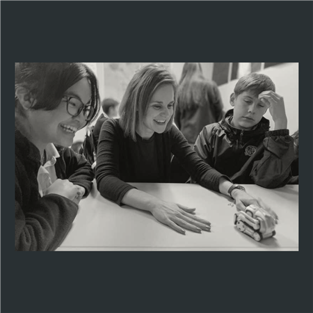 Rebecca Luebker laughing at a table with two students as they watch a robot move around